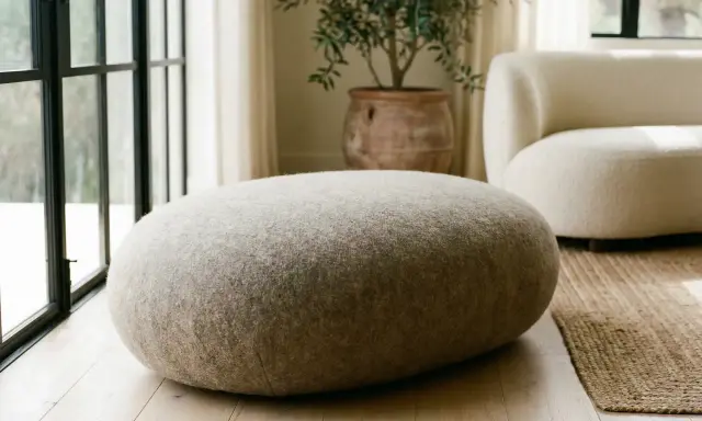 Un morbido pouf dal design organico, rivestito in tessuto grezzo, si abbina a un divano bouclé e a un vaso in terracotta.