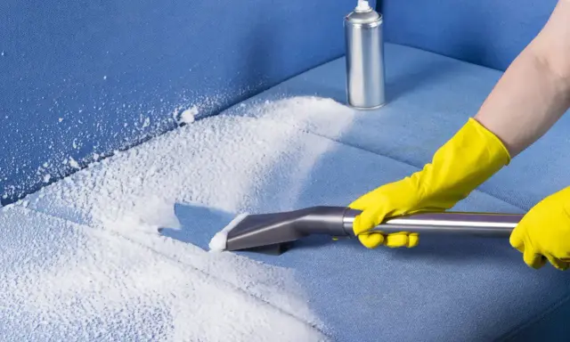 Mani con guanti gialli usano un aspiratore per pulire un divano in microfibra con schiuma detergente.