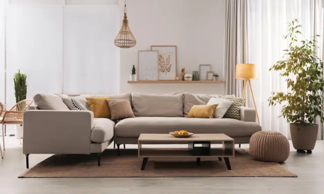 Divano angolare color taupe, con cuscini decorativi, tavolino in legno e pianta verde.