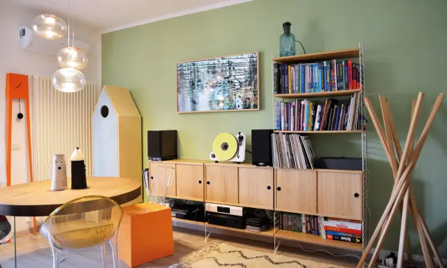 Soggiorno con pareti verde salvia, mobili in legno chiaro, giradischi giallo e un quadro astratto. I colori per pareti soggiorno sono rilassanti.