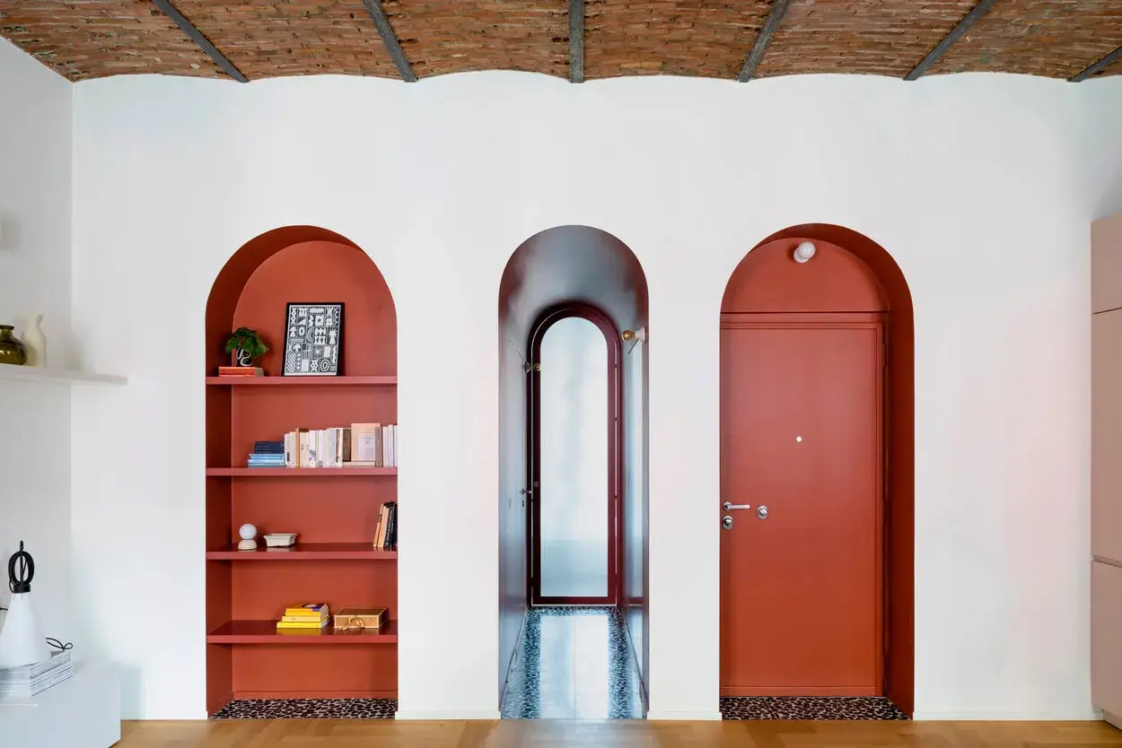 Una nicchia nel muro con mensole color terracotta ospita libri e decorazioni, accanto a un corridoio e una porta rossa.
