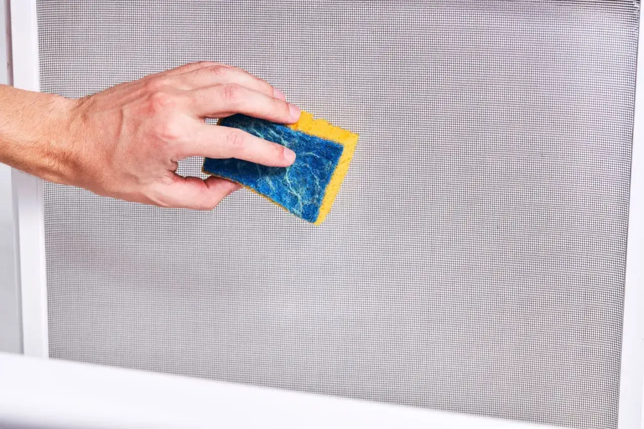Mano che pulisce una zanzariera con una spugna blu e gialla. Un modo semplice per imparare come pulire le zanzariere.