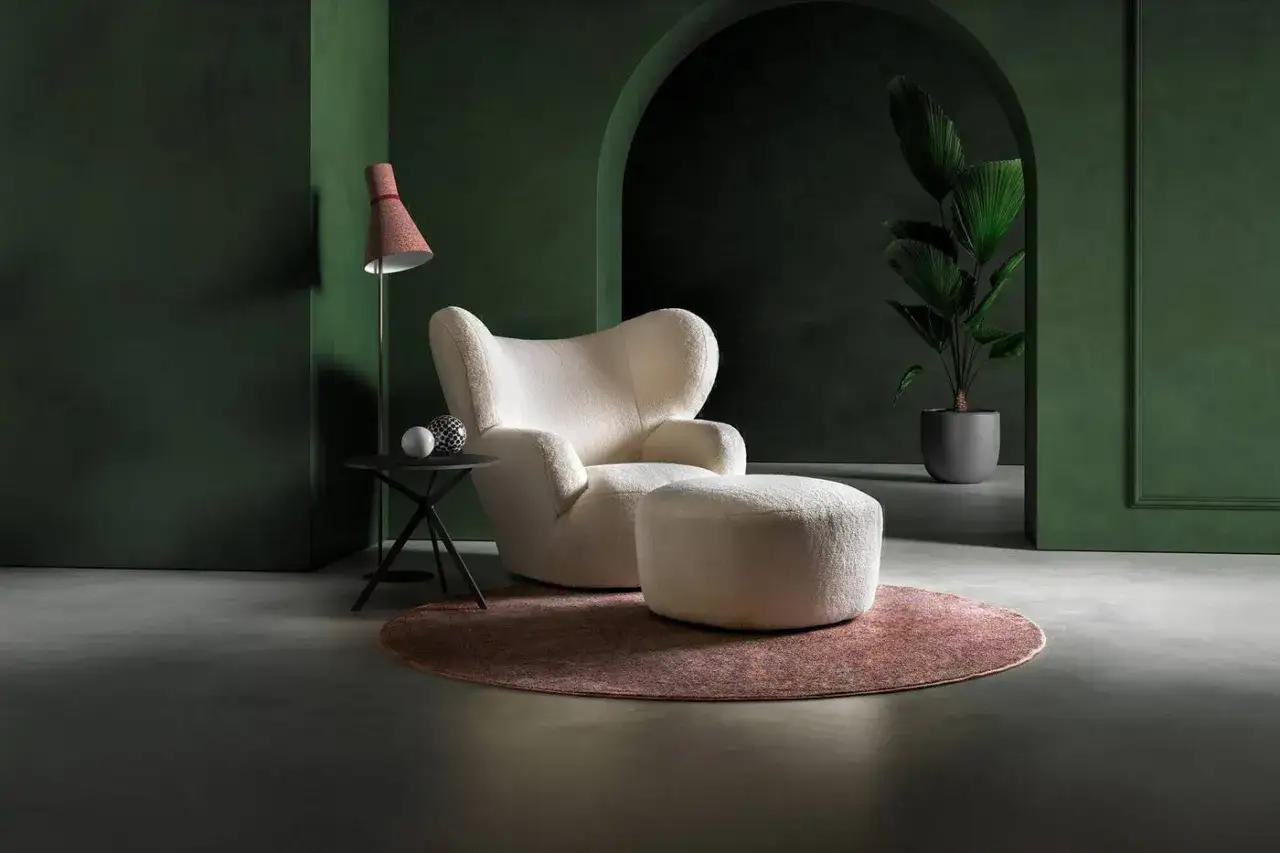 Poltrona avvolgente color crema con pouf, tappeto rotondo, lampada da terra e pianta in vaso. Un'atmosfera di puro adrenalina design.