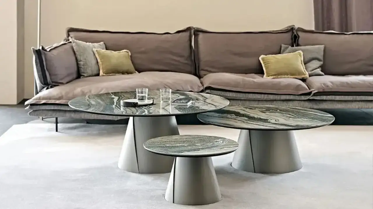 Un elegante tavolino design con piano in marmo verde e base conica grigia, accanto a un divano moderno.