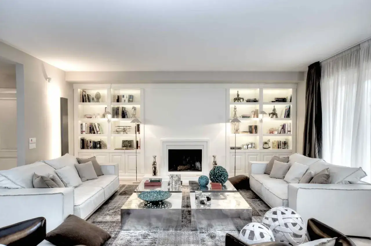 Elegante soggiorno beige e bianco con divani, librerie illuminate, camino e tavolini metallici.