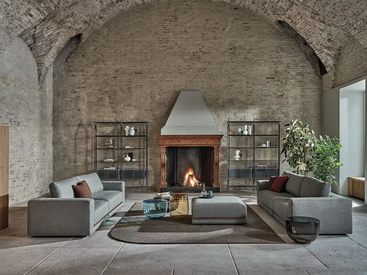 Elegante salone con camino in pietra, divani grigi, tavolini in vetro e librerie. Un esempio di come arredare un salone moderno in un contesto rustico.