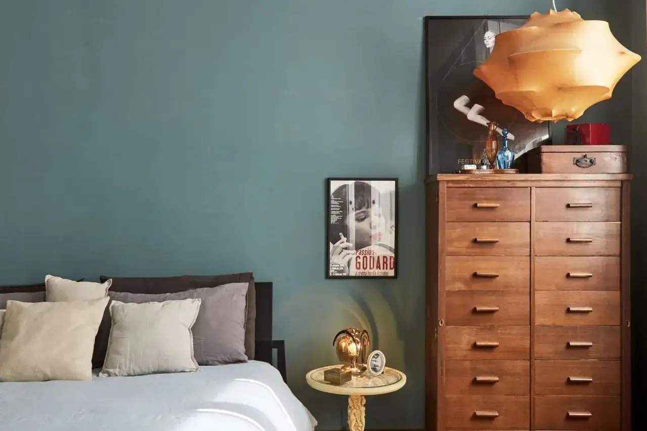Camera da letto con pareti color ottanio, un com&ograve; in legno scuro e un letto con cuscini in tonalit&agrave; neutre. L'abbinamento colori crea un'atmosfera rilassante.