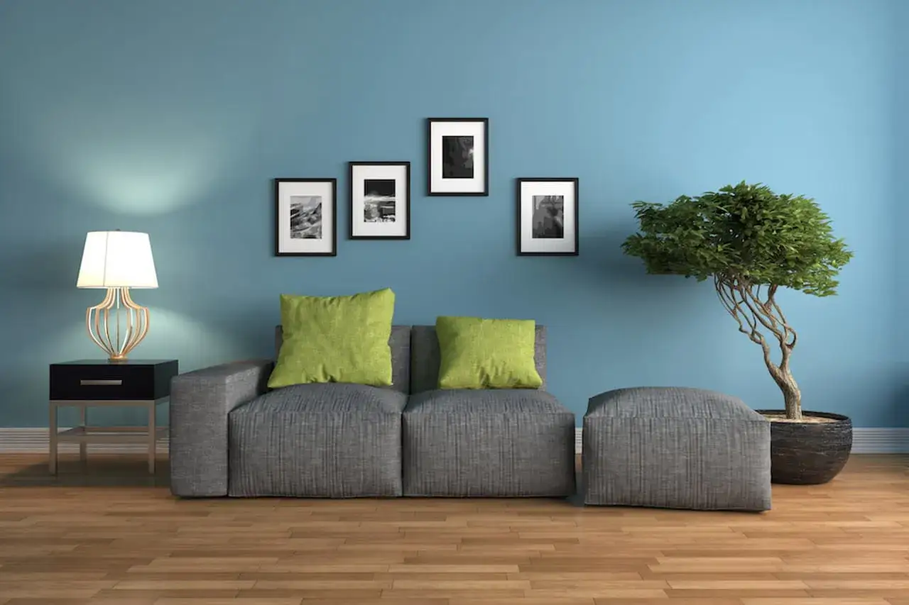 Soggiorno con divano grigio, cuscini verdi, quadri astratti e un bonsai su sfondo blu con colori freddi.