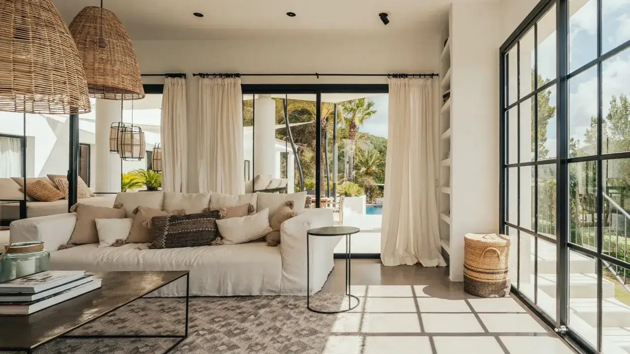 Soggiorno luminoso con divano bianco, cuscini in lino e rattan. Idee originali per arredare casa al mare: lampade in vimini e vista su piscina.