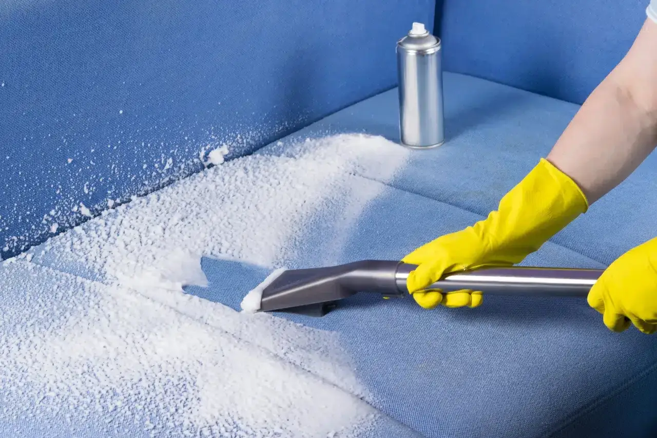 Mani con guanti gialli usano un aspiratore per pulire un divano blu, applicando schiuma detergente. Guida su come pulire divano alcantara.