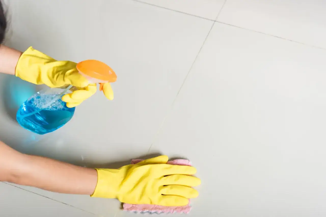 Mani con guanti gialli spruzzano detergente blu su piastrelle chiare e puliscono con un panno. Ecco come pulire le piastrelle del bagno.
