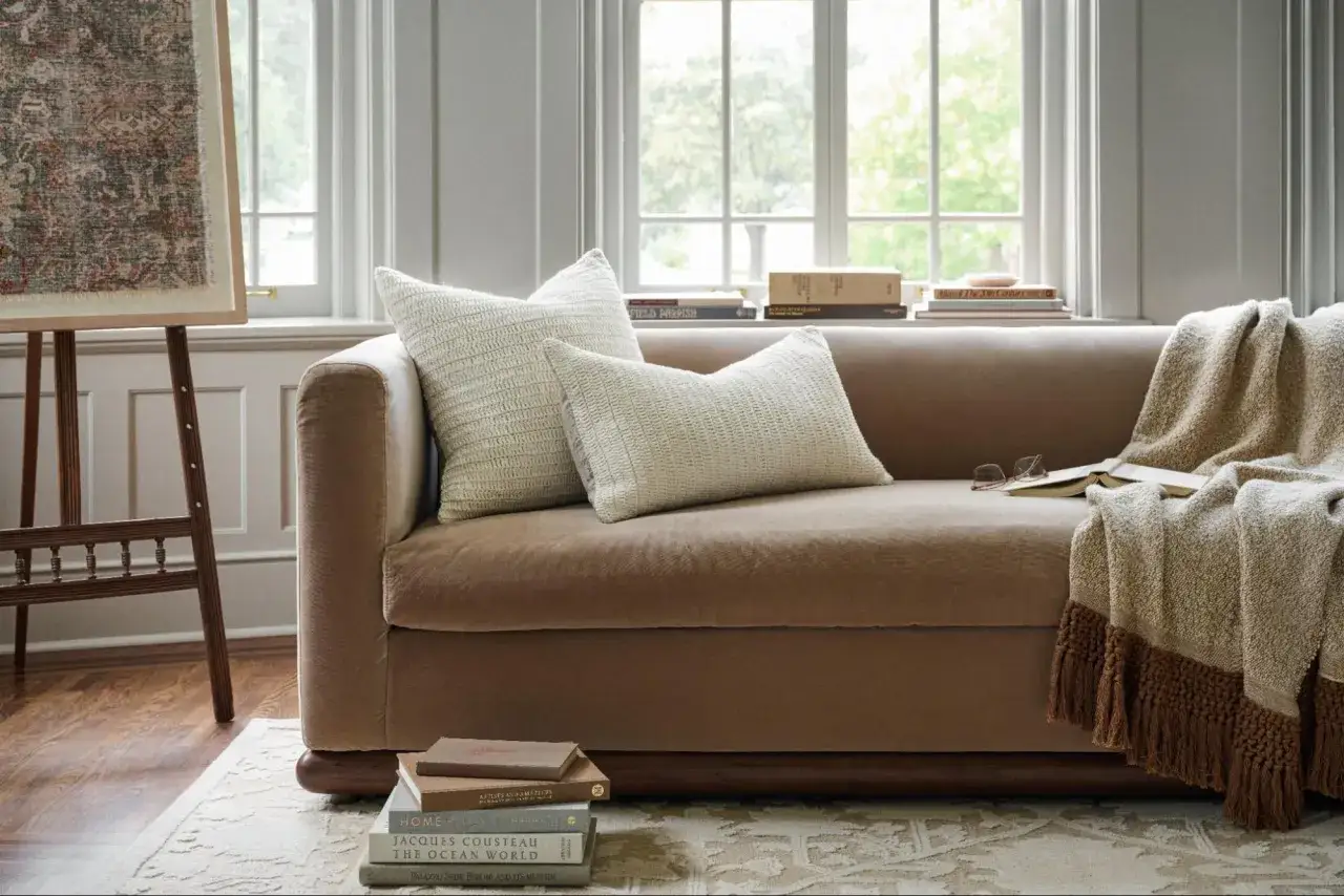 Un divano marrone con una morbida texture, cuscini decorativi e una coperta con frange. Libri e occhiali appoggiati sul bracciolo.