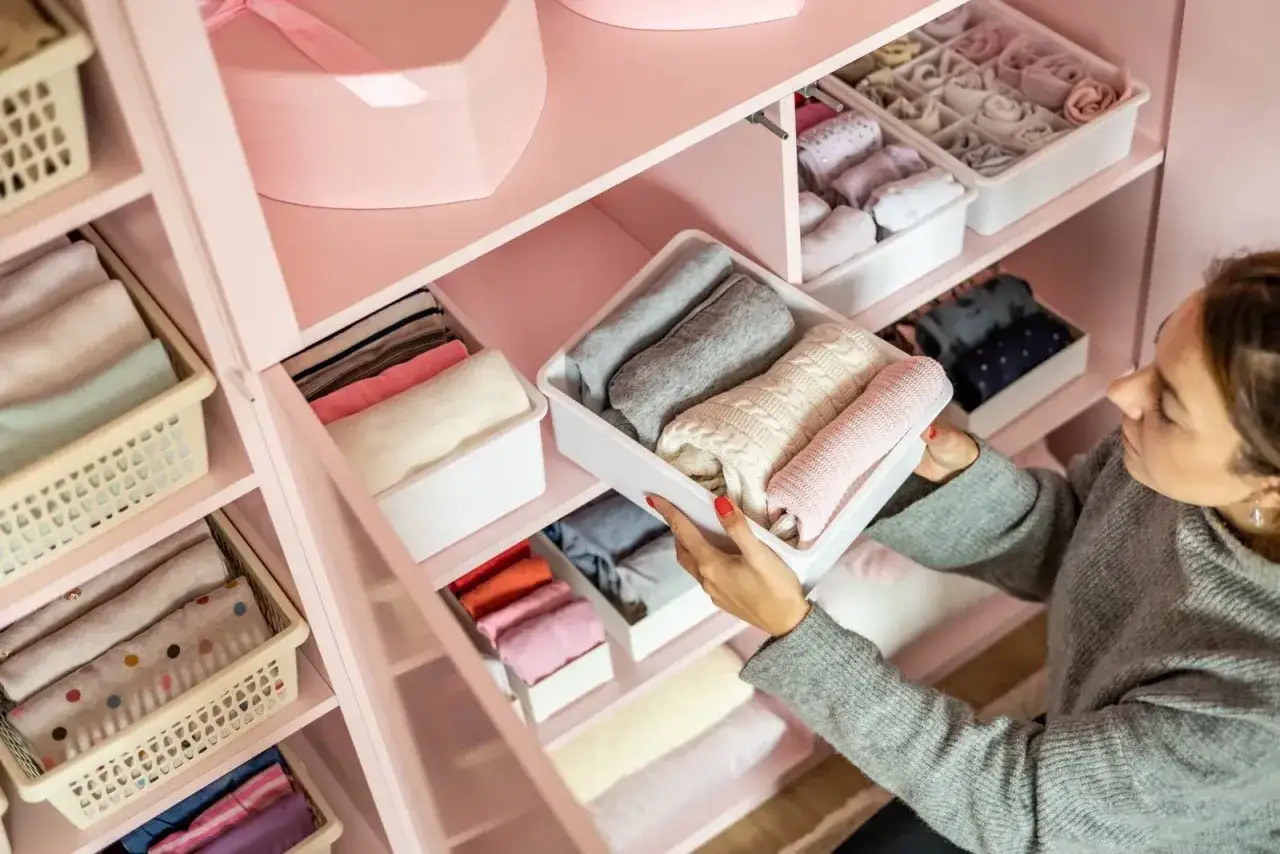 Donna sistema maglioni piegati in contenitori bianchi, un esempio di come fare decluttering in modo ordinato e funzionale.