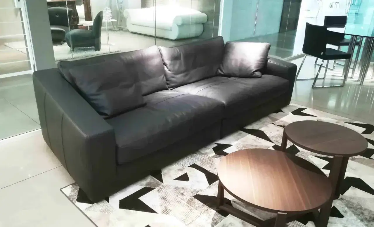 Un elegante divano angolare in pelle nera, con due tavolini rotondi in legno, su un tappeto geometrico. Un frighetto è visibile in lontananza.