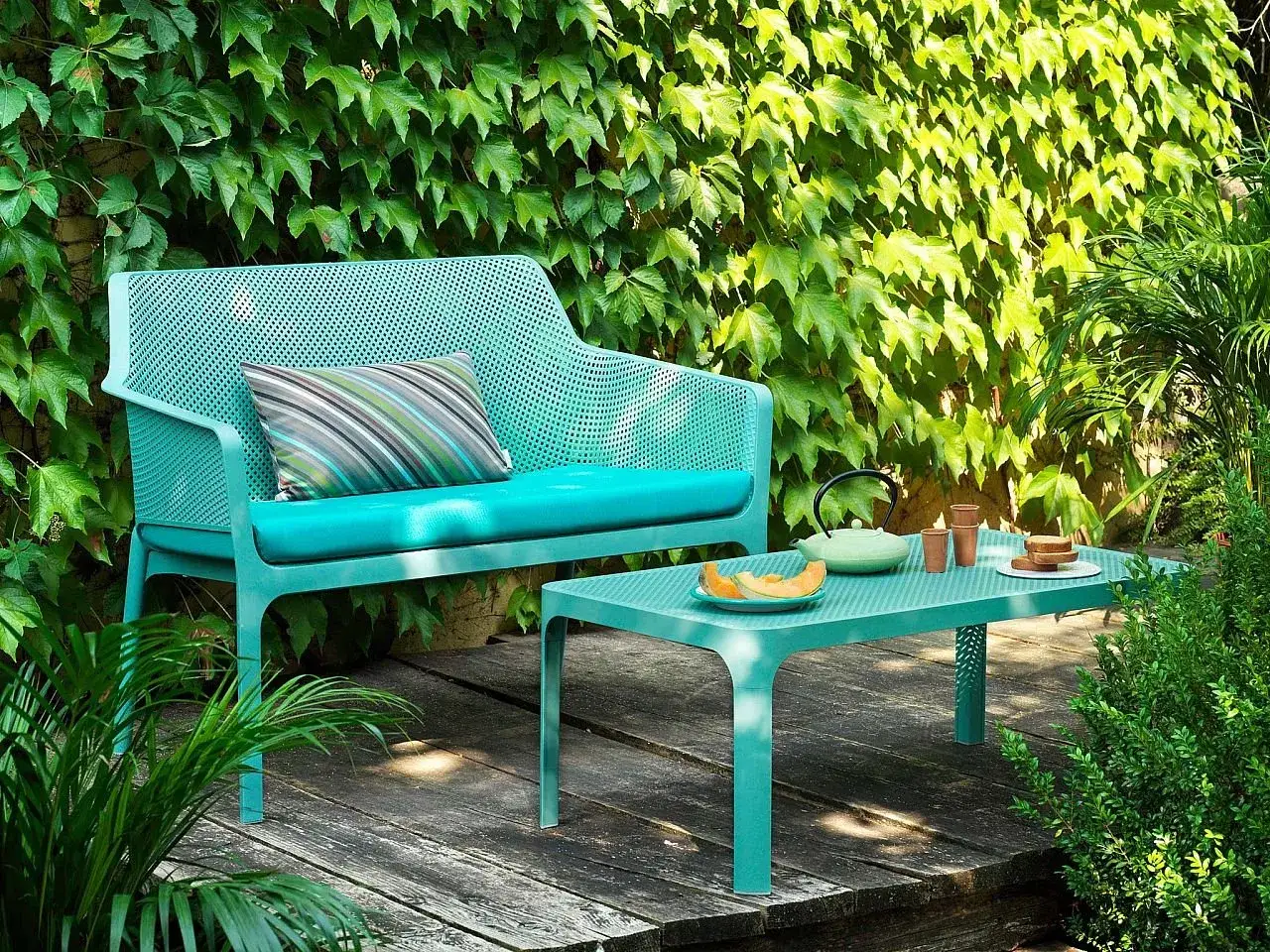 Divano e tavolino Nardi Spa Chiampo in un giardino rigoglioso, con cuscini color acqua e un set da t&egrave;.