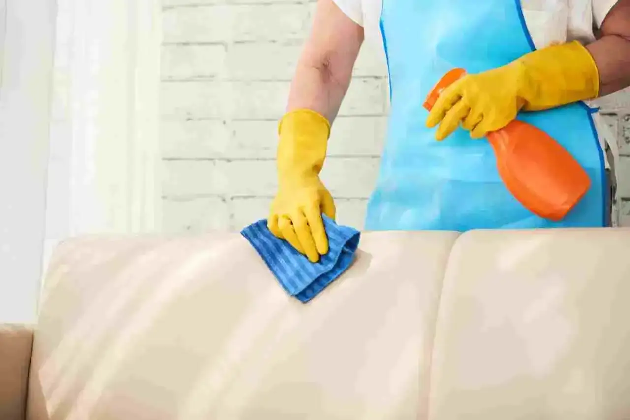 Mani con guanti gialli puliscono un divano non sfoderabile che puzza con uno spray e un panno blu.