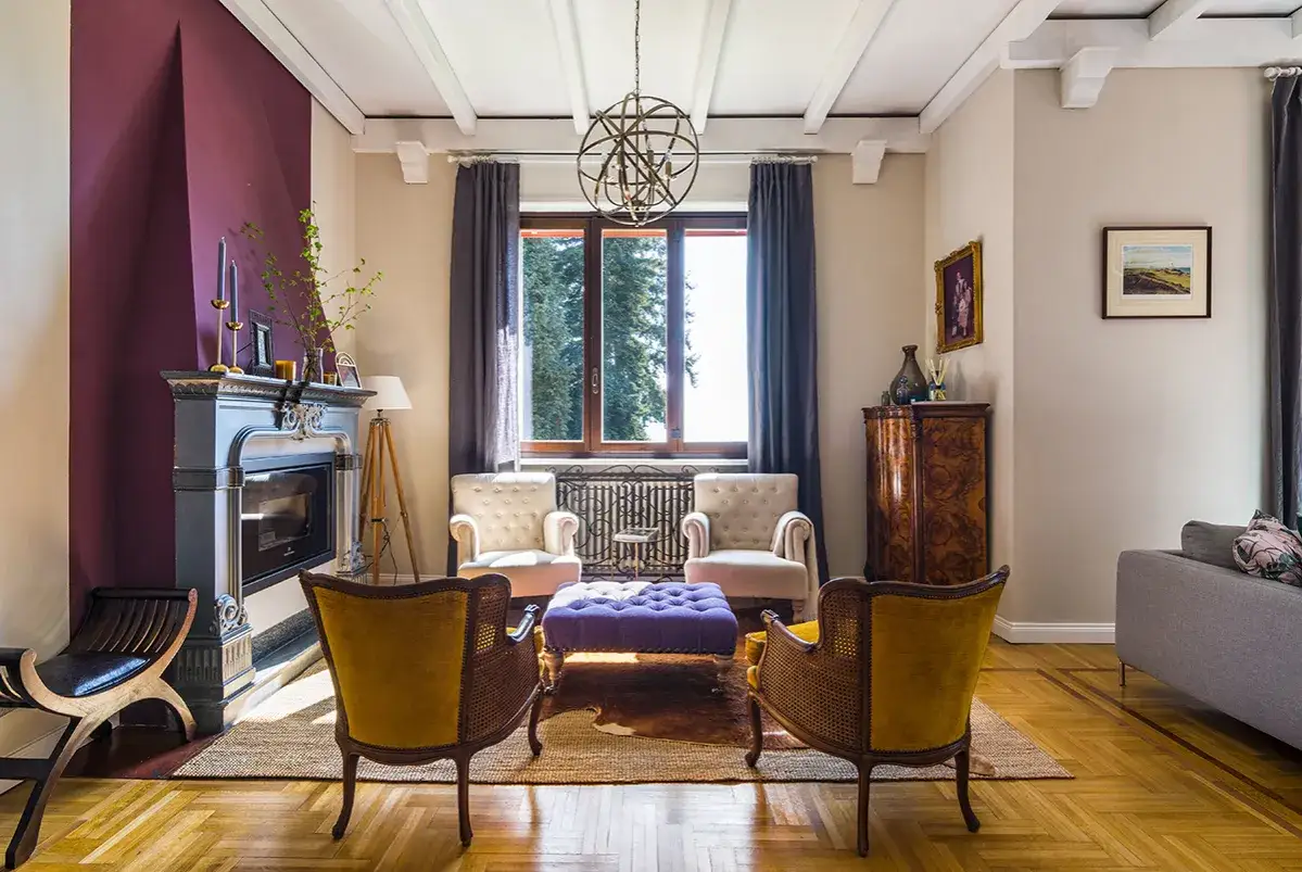 Salotto elegante con camino, poltrone vintage e un pouf viola. Un mix di stili per idee arredo uniche.