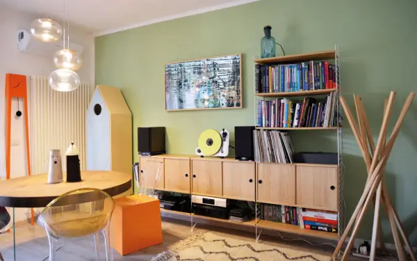 Soggiorno con pareti verde salvia, mobili in legno chiaro, giradischi giallo e un quadro astratto. I colori per pareti soggiorno sono rilassanti.