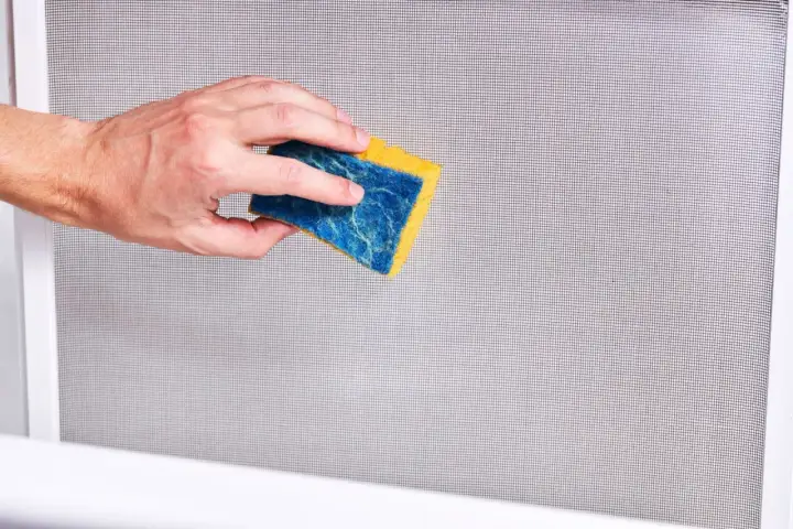 Mano che pulisce una zanzariera con una spugna blu e gialla. Un modo semplice per imparare come pulire le zanzariere.