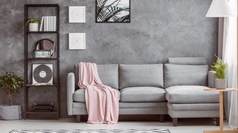 Un divano grigio con coperta rosa, una libreria nera con piante e vinile, e un tavolino di legno: ecco cosa mettere vicino al divano per un tocco di stile.