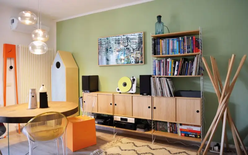 Soggiorno con pareti verde salvia, mobili in legno chiaro, giradischi giallo e un quadro astratto. I colori per pareti soggiorno sono rilassanti.